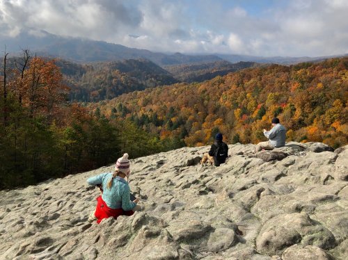 Three people explore a rocky outcrop with a colorful autumn landscape and mountains in the background.