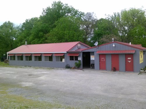A gray building with a red roof, featuring large windows and a red door, surrounded by greenery.