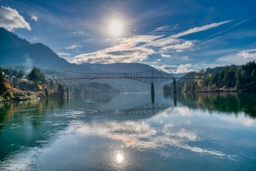 A serene river scene with a bridge, mountains, and a bright sun reflecting on the water under a blue sky.