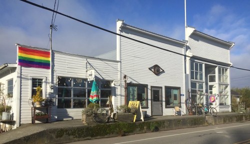 A charming white building with large windows, featuring a rainbow flag and outdoor decor, set against a blue sky.