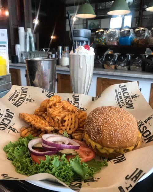 A plate with a cheeseburger, curly fries, lettuce, tomato, and onion, alongside a milkshake in a diner setting.