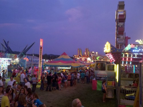 A vibrant carnival scene at dusk, featuring colorful rides, tents, and crowds enjoying the festivities.