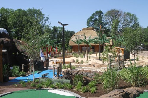 A mini-golf course featuring tropical landscaping, palm trees, and a thatched-roof building under a clear blue sky.