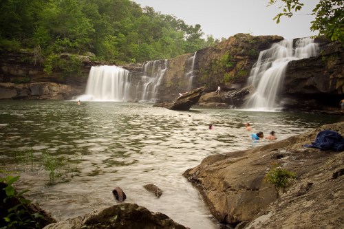 A serene waterfall cascades into a calm pool, with people swimming and enjoying the lush, green surroundings.