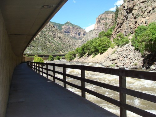 A scenic view of a river flowing beside a walkway, flanked by rocky cliffs and lush greenery under a blue sky.