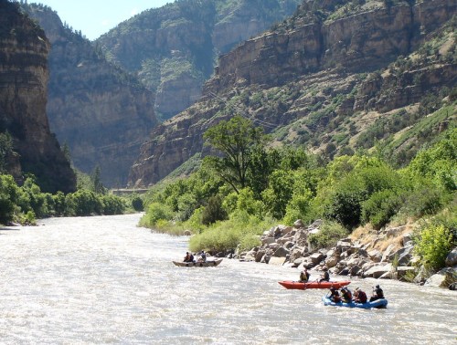 A river flows through a canyon, with kayakers navigating the water surrounded by lush greenery and rocky cliffs.