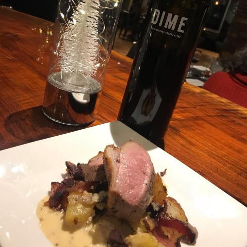 A plate of sliced pork with bacon and potatoes, next to a decorative tree and a bottle of DIME wine.