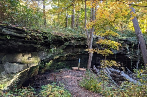 A rocky overhang in a forest, surrounded by trees with autumn foliage and a small trail leading into the woods.
