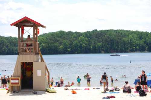 A busy beach scene with a lifeguard tower, people swimming, and a forested shoreline in the background.