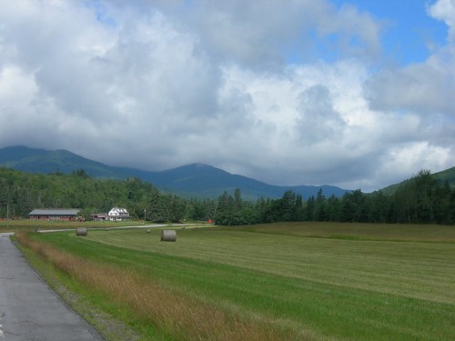 A scenic landscape featuring green fields, hay bales, and mountains under a partly cloudy sky.