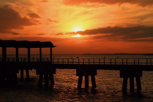 Silhouette of a pier against a vibrant orange sunset over calm waters.