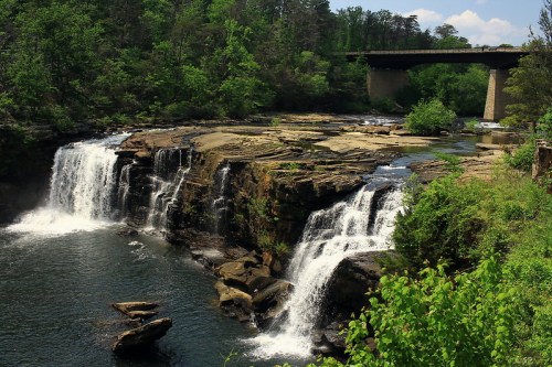 A scenic view of a waterfall cascading over rocks, surrounded by lush greenery and a bridge in the background.