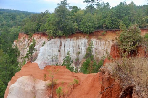 A scenic view of layered red and white cliffs surrounded by lush green trees under a cloudy sky.
