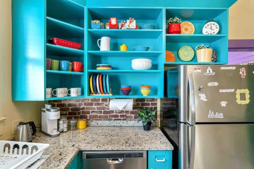 Brightly colored kitchen with turquoise shelves, various dishes, and a stainless steel fridge against a brick wall.