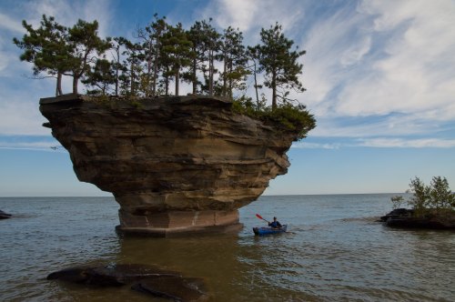 A kayaker paddles near a unique rock formation topped with trees, surrounded by calm water and a blue sky.