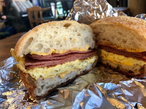 A close-up of a sandwich with layers of ham and scrambled eggs on toasted bread, wrapped in foil.