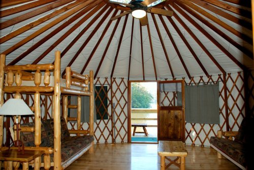 Interior of a cozy yurt with wooden beams, bunk beds, a lamp, and seating, leading to an outdoor view.