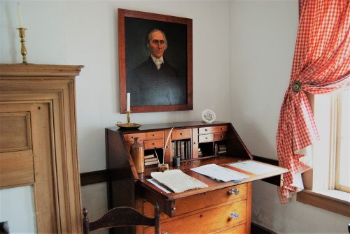 A vintage desk with papers and a quill, beside a portrait and a window with checkered curtains.