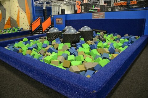 A colorful foam pit filled with green, blue, and gray blocks, surrounded by safety padding in a trampoline park.
