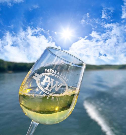 A glass of white wine with a winery logo, set against a bright sky and serene lake backdrop.
