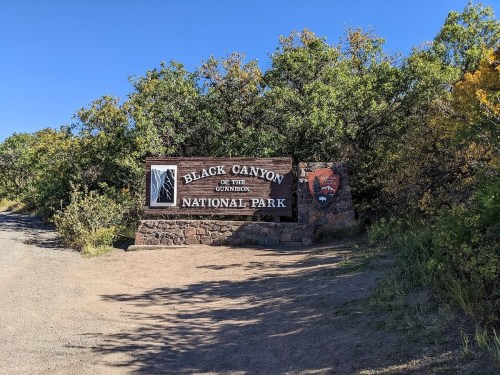 Sign for Black Canyon of the Gunnison National Park, surrounded by greenery and a clear blue sky.