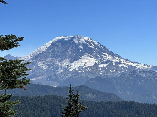 A snow-capped mountain rises above a forested landscape under a clear blue sky.