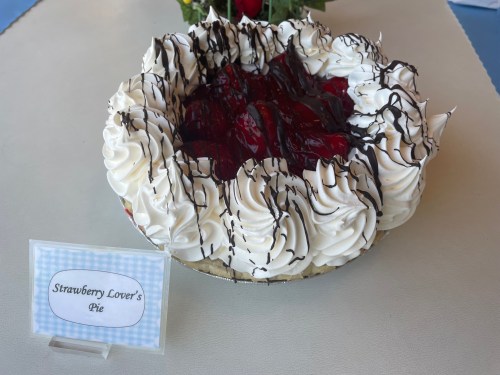 A pie topped with whipped cream, strawberries, and chocolate drizzle, labeled "Strawberry Lover's Pie."