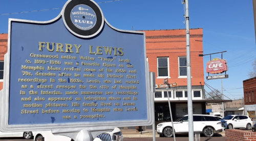 Historical marker about Furry Lewis, a key figure in the Memphis blues scene, with a café and buildings in the background.