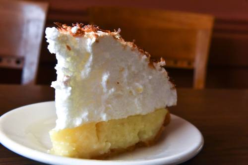 A slice of coconut cream pie on a white plate, featuring a fluffy meringue topping and a yellow custard filling.