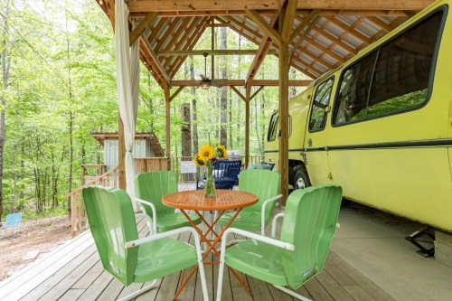 A cozy outdoor seating area with a round table and green chairs, surrounded by trees and a yellow van nearby.