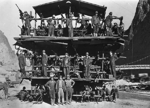 Historical black and white photo of workers posing on a large construction platform in a mountainous area.