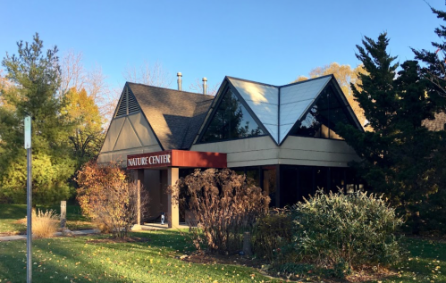 A modern nature center building with a triangular roof, surrounded by trees and shrubs in a sunny outdoor setting.