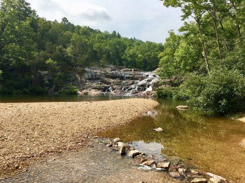 A serene landscape featuring a waterfall cascading into a calm pool, surrounded by lush green trees and rocky terrain.