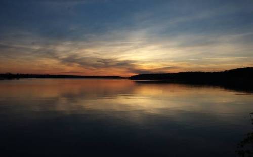 A serene lake at sunset, reflecting colorful clouds and a calm sky, surrounded by trees on the horizon.