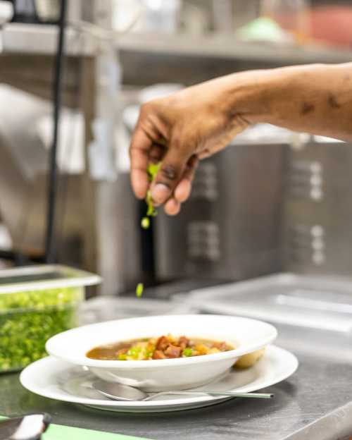 A hand sprinkles fresh herbs over a bowl of soup in a kitchen setting.