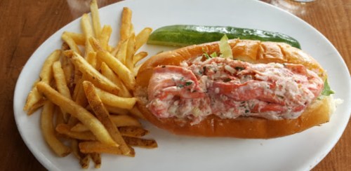 A lobster roll with lettuce on a bun, served with a pickle and a side of golden French fries.