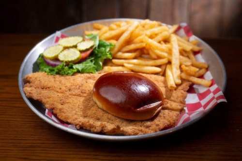 A plate featuring a large fried fish fillet, a bun, fries, and a side of pickles and lettuce.