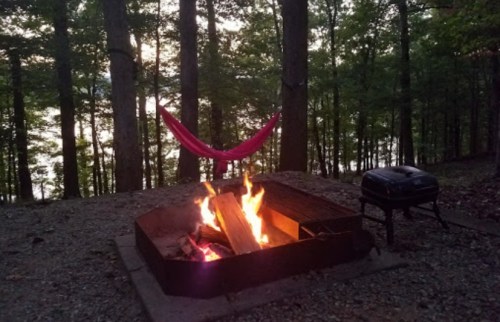 A cozy campsite with a fire pit, burning logs, a grill, and a hammock set among trees by a lake at sunset.