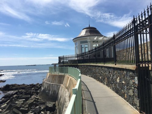 A scenic walkway along the coast, featuring a gazebo and rocky shoreline under a clear blue sky.