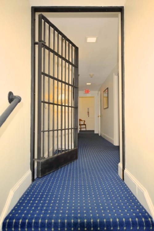 A hallway with a blue carpet and a barred door partially open, leading to a well-lit area beyond.