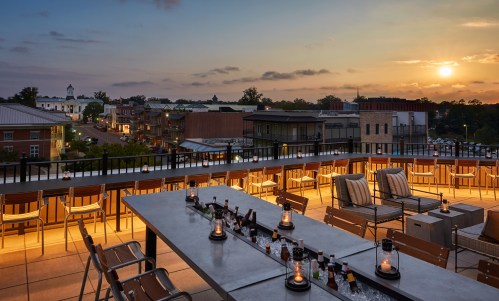 Rooftop terrace with a long table, candles, and a sunset view over a town with buildings and trees in the background.