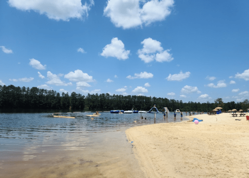 A sunny beach scene with clear water, sandy shore, and people enjoying various activities under a blue sky with clouds.