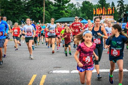 A group of runners, including children and adults, participating in a race on a street lined with trees and flags.