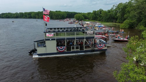 A floating restaurant on a river, surrounded by boats and greenery, with flags and festive decorations.