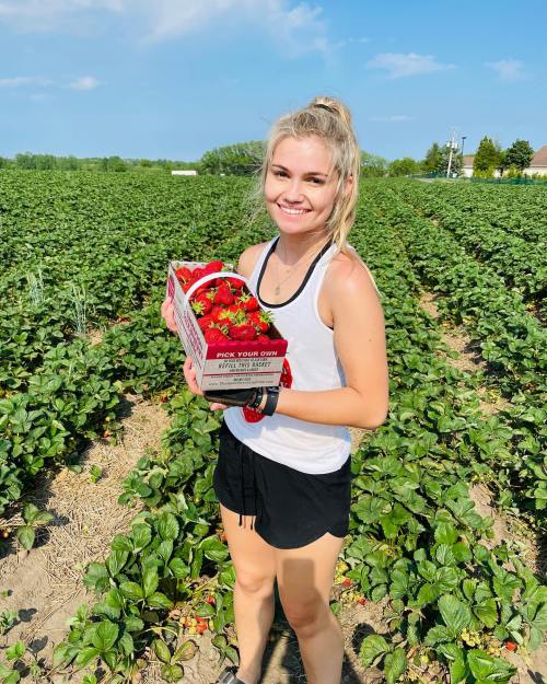 A smiling young woman holds a box of strawberries in a lush green field.