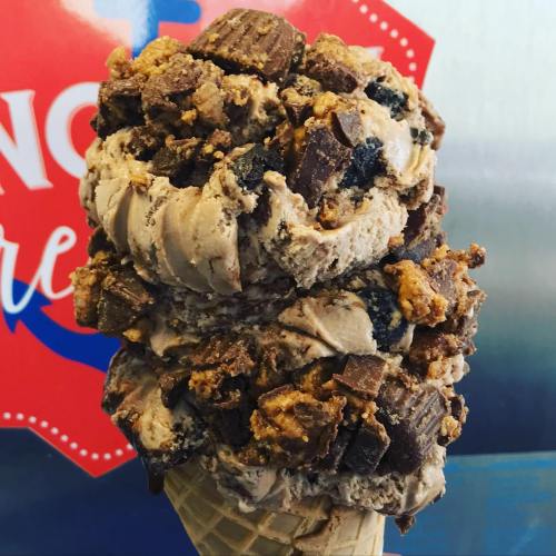 A large ice cream cone with two scoops of chocolate ice cream topped with chunks of cookie and candy.