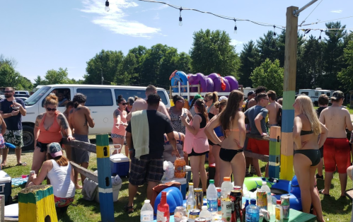 A lively outdoor gathering with people in swimsuits, food, drinks, and a van in the background on a sunny day.