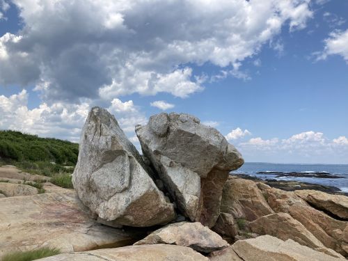 A large split rock on a coastal landscape under a partly cloudy sky, with the ocean visible in the background.