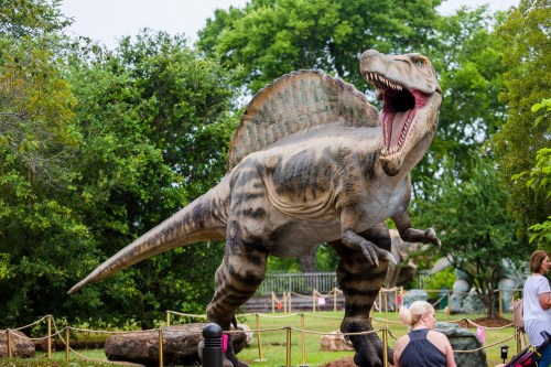 A large, roaring dinosaur statue with a spiked back, surrounded by greenery and visitors in a park setting.