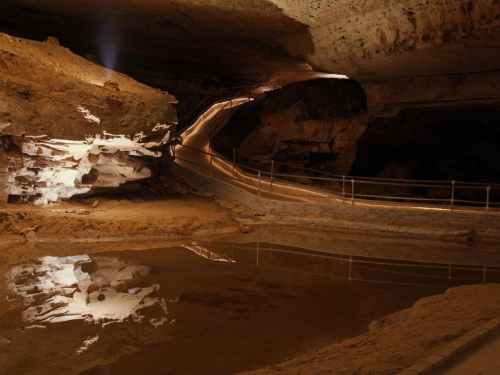 A dimly lit cave with rock formations, a winding path, and a reflective pool of water.